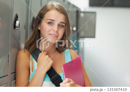 Teenage student standing in school hallway by gray lockers carrying pink binder and backpack strap 135307410