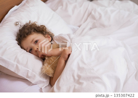 Girl with curly hair wearing pajamas hugging teddy bear lying on bed in bedroom, copy space 135307422