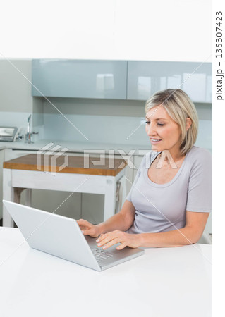 Woman typing on laptop at home kitchen table with chopping block, cabinets and faucet, copy space 135307423