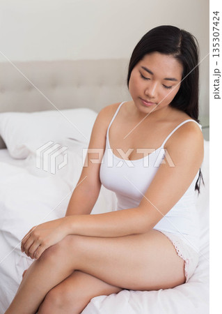 Asian woman sitting on bed wearing white camisole and pink lace shorts in bedroom, copy space Asian woman sitting on bed wearing white camisole and pink lace shorts in bedroom, copy space 135307424