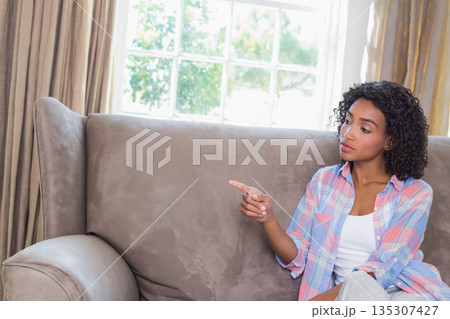African American woman sitting on sofa pointing left at home window with beige curtains, copy space African American woman sitting on sofa pointing left at home window with beige curtains, copy space 135307427
