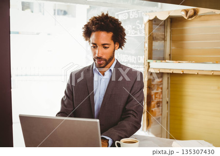 African American man wearing dark suit typing on laptop at coffee shop with coffee cup 135307430