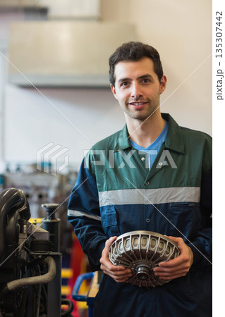Man in coverall holding component by engine stand in workshop near overhead hood, copy space 135307442