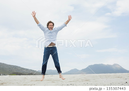 Mid adult man jumping on sandy shoreline wearing light blue shirt and dark jeans barefoot Mid adult man jumping on sandy shoreline wearing light blue shirt and dark jeans barefoot 135307443