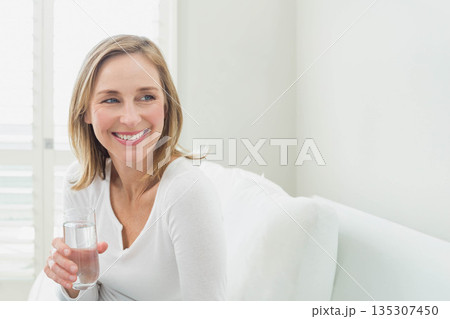 Senior woman sitting on white sofa in bright living room holding glass of water, copy space 135307450