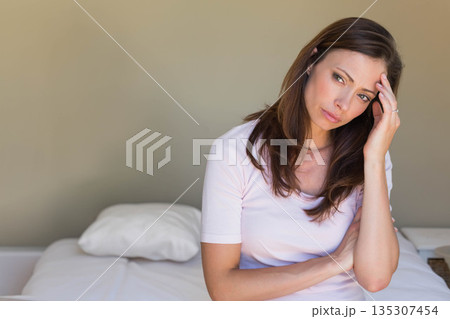 Woman sitting on bed edge touching temple near white pillow and woven basket, copy space 135307454
