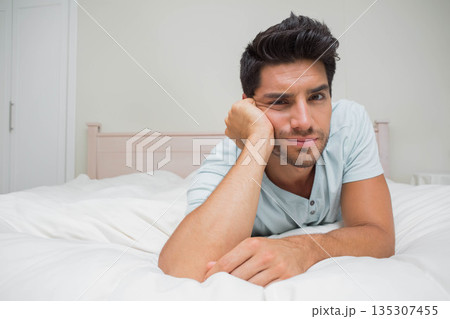 Male lying on bed with white comforter, head resting on hand, gazing toward camera in bedroom 135307455