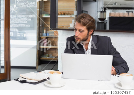 Man wearing dark suit typing on laptop at café table, writing in notebook beside coffee cups Man wearing dark suit typing on laptop at café table, writing in notebook beside coffee cups 135307457