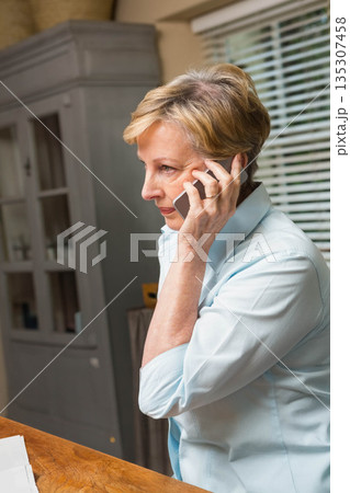 Senior woman in blue shirt holding smartphone to ear at kitchen table with papers, copy space 135307458