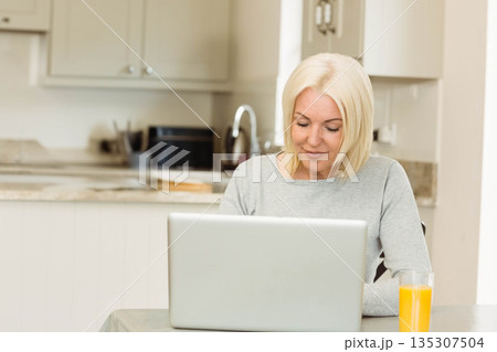 Woman browsing on laptop in modern kitchen while sipping orange juice beside sink Woman browsing on laptop in modern kitchen while sipping orange juice beside sink 135307504