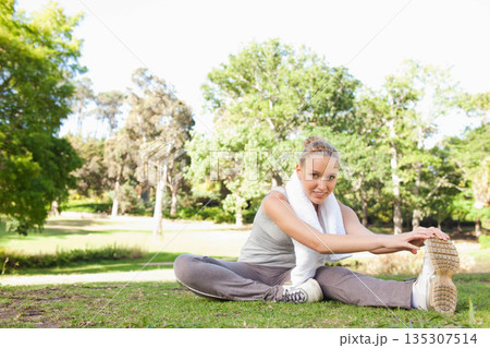 Woman straddling grassy lawn in park wearing grey sportswear with white towel and shoes 135307514