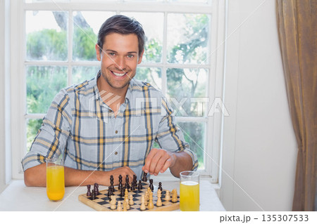 Middle-aged man sitting at dining table by window moving chess pieces near glasses of orange juice 135307533