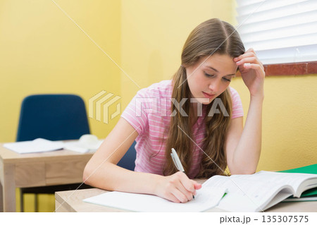 Teenage female student writing in notebook with pen at wooden desk in study room with textbook Teenage female student writing in notebook with pen at wooden desk in study room with textbook 135307575