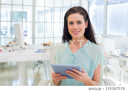 Female professional in light green blouse holding tablet and sitting amid desks, monitors in office Female professional in light green blouse holding tablet and sitting amid desks, monitors in office 135307578