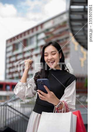 Asian woman office worker standing carrying shopping bags and making a fist while looking at phone. Asian woman office worker standing carrying shopping bags and making a fist while looking at phone. 135307861