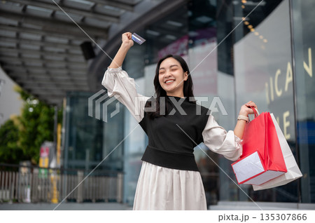 Asian woman female office worker standing outside mall carrying shopping bag and raising credit card 135307866