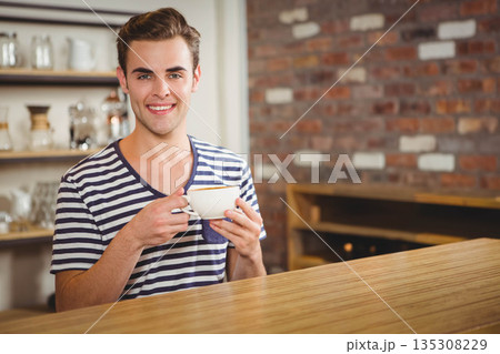 Mid adult man holding white ceramic coffee cup behind counter in cafe with exposed brick wall Mid adult man holding white ceramic coffee cup behind counter in cafe with exposed brick wall 135308229
