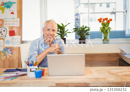 Senior man sitting at desk in office, smiling and typing on silver laptop beside colored pencils 135308232