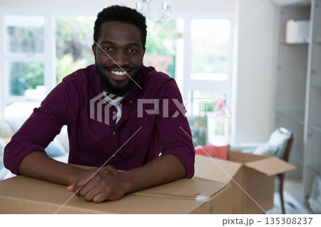 African American man leaning on cardboard box under chandelier at home surrounded by moving boxes 135308237