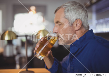 Senior man lifting pale beer glass at wooden bar counter under brass lamp, copy space 135308245