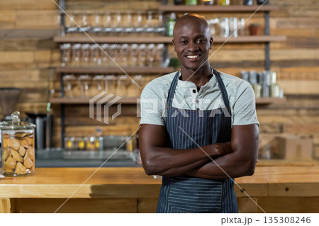 Standing African American man wearing apron behind bar counter in cafe with glass jar cookies Standing African American man wearing apron behind bar counter in cafe with glass jar cookies 135308246