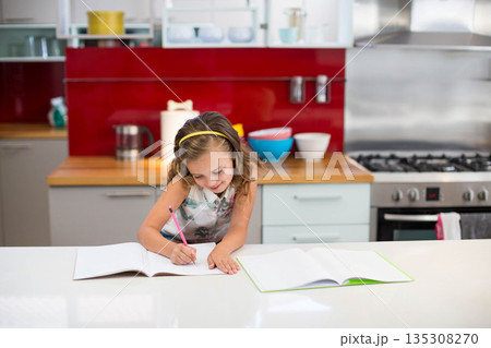 School-age girl writing in notebook at kitchen island with colorful mixing bowls, metal kettle 135308270