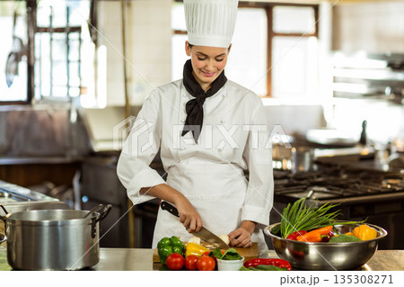 Female chef slicing onion with chef's knife on wooden cutting board at commercial kitchen prep area 135308271