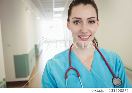 Female nurse standing in hospital corridor wearing light blue scrubs and red stethoscope 135308272