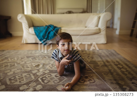 Boy lying on patterned rug in living room holding remote control and watching TV 135308273