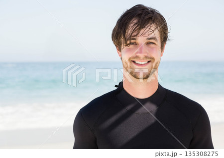 Man standing near waterline on sandy beach wearing black wetsuit and smiling, copy space 135308275