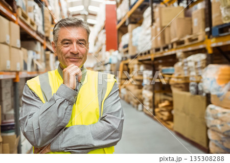 Senior male standing in warehouse aisle inspecting inventory on racks wearing safety vest 135308288