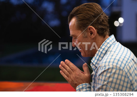 Senior man praying at patio table by red tablecloth and pool under porch lights, copy space Senior man praying at patio table by red tablecloth and pool under porch lights, copy space 135308294