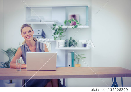 Woman smiling while sitting at wooden desk with laptop, water glass, plants, binders and folders Woman smiling while sitting at wooden desk with laptop, water glass, plants, binders and folders 135308299