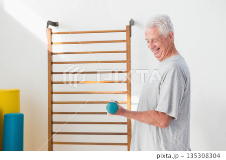 Senior man lifting teal hand weight beside exercise ladder with foam rollers in gym, copy space Senior man lifting teal hand weight beside exercise ladder with foam rollers in gym, copy space 135308304