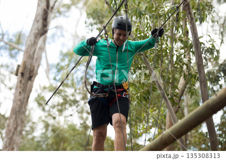 Man balancing on suspended wooden beam in forest, gripping steel cables with helmet and harness 135308313