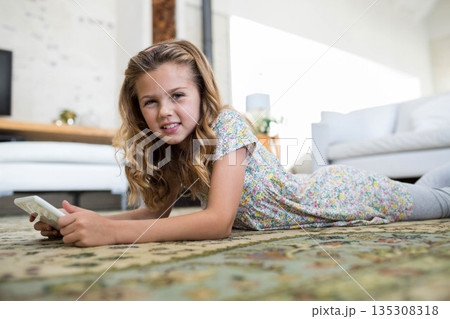 Female child lying on patterned rug in bright living room holding white smartphone 135308318