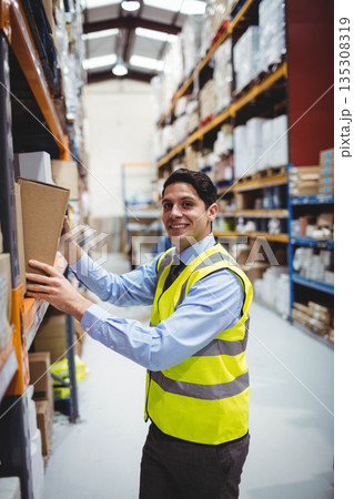 Male worker wearing safety vest reaching to retrieve cardboard box from shelving in storage aisle Male worker wearing safety vest reaching to retrieve cardboard box from shelving in storage aisle 135308319
