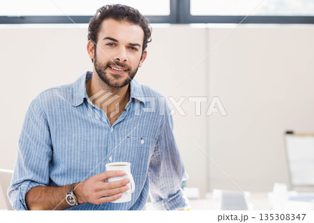 Adult man sitting at desk in bright office wearing wristwatch and holding white ceramic coffee mug 135308347