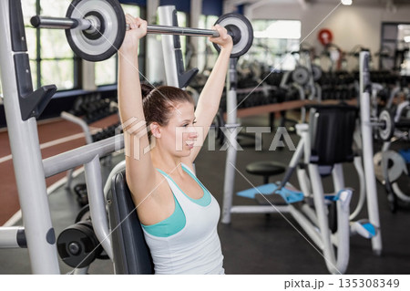 Female gym-goer pressing barbell overhead on padded weight machine bench in gym training area 135308349