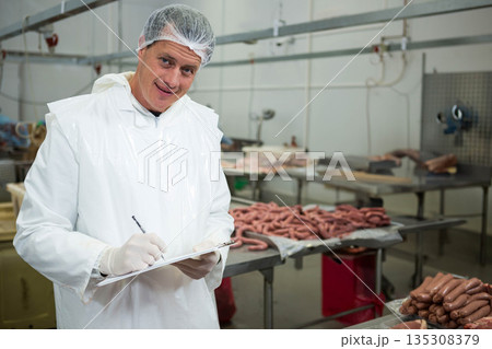 Male inspector wearing coat examining sausage trays at meat plant holding clipboard, copy space Male inspector wearing coat examining sausage trays at meat plant holding clipboard, copy space 135308379