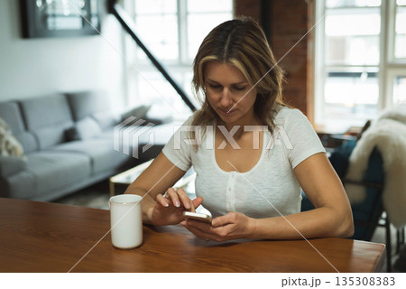 Woman holding smartphone while sitting at wooden table with ceramic mug in loft living room Woman holding smartphone while sitting at wooden table with ceramic mug in loft living room 135308383