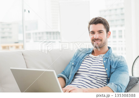 Man working on laptop while sitting on light-colored sofa near floor-to-ceiling window, copy space Man working on laptop while sitting on light-colored sofa near floor-to-ceiling window, copy space 135308388