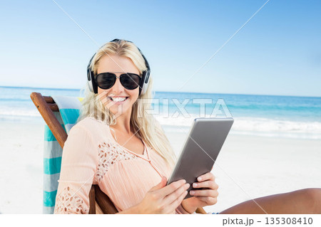 Blonde woman resting on towel-draped lounger using tablet wearing sunglasses, headphones on beach Blonde woman resting on towel-draped lounger using tablet wearing sunglasses, headphones on beach 135308410