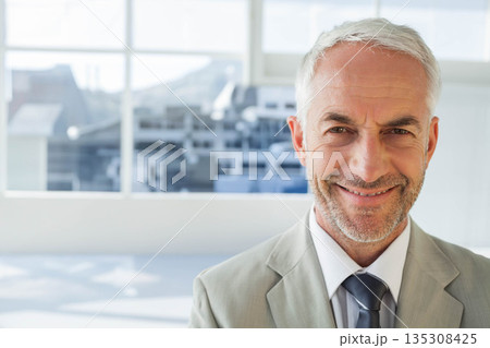 Senior man wearing suit smiling while standing in office by windows overlooking skyline, copy space Senior man wearing suit smiling while standing in office by windows overlooking skyline, copy space 135308425