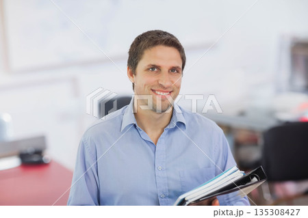 Man wearing light blue shirt standing in bright office space holding black ring binder and papers 135308427
