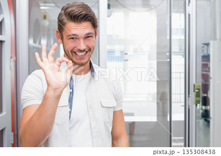 Man standing before glass partition and sliding door in modern office, raising right hand OK sign 135308438