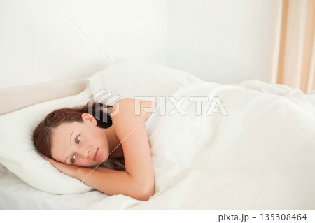 Woman lying on side wearing white tank top in bed with pillow and duvet, copy space 135308464