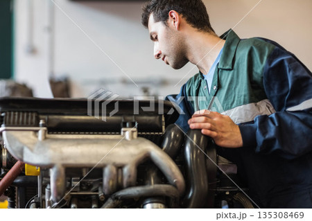 Man wearing two-tone coverall leaning over engine inspecting manifold hose in garage, copy space 135308469