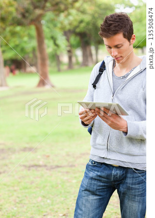 Man standing on grassy lawn in park using tablet and carrying backpack, copy space 135308474