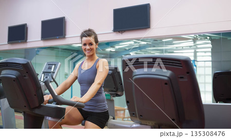 Woman smiling at camera while pedaling on stationary bike in gym with mirrored wall and TVs 135308476